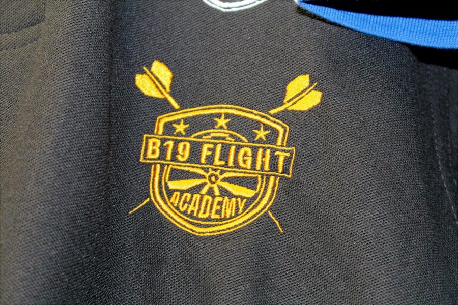 Close-up of B19 Flight Academy darts academy logo embroidered in gold thread on a black polo shirt
